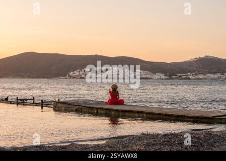 Eine Person in rotem Kleid sitzt auf einem hölzernen Pier am Papikinou Beach, Milos, Griechenland, und bestaunt den ruhigen Sonnenuntergang über einer malerischen Stadt und den Hügeln in der Stockfoto