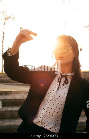 Eine Frau in Sonnenbrille und einer Polka Dot-Bluse, umrahmt von Sonnenlicht, hebt ihre Hand, während sie draußen steht, und schafft ein ausdrucksstarkes und dynamisches Bild Stockfoto