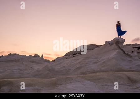 Rückansicht einer nicht erkennbaren Frau in einem fließenden blauen Kleid, die auf den glatten weißen Felsformationen des Sarakiniko Beach auf der Insel Milos, G, steht Stockfoto