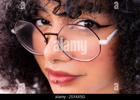 Nahaufnahme einer stilvollen lateinfrau mit großer Brille, lockigem Haar und Gesichtspiercings, die einen ausdrucksstarken und selbstbewussten Look zeigt. Einfängt moderne B Stockfoto