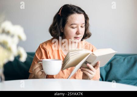 Die junge Frau liest aufmerksam, während sie eine Tasse Kaffee hält, bequem auf einem Sofa zu Hause sitzt und einen friedlichen Moment in der Wohnung verkörpert. Stockfoto