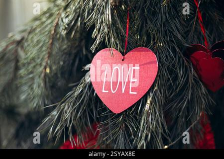 Rotes herzförmiges Papier mit LIEBE geschrieben, hängt an einem immergrünen Baumzweig. Perfekt, um die Essenz der Romantik und des Valentinstages festzuhalten. Stockfoto