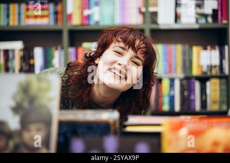 Porträt einer fröhlichen jungen Frau mit lockigen Haaren, die in der Nähe eines Bücherregals mit bunten Büchern lächelt, in einer Bibliothek. Stockfoto