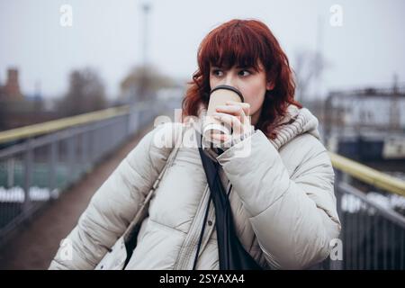 Eine junge Frau in einem warmen Wintermantel trinkt an einem Tag mit dem Zug Kaffee aus einer Einwegtasse, die auf einem Weg mit verschwommenem Hintergrund steht. Stockfoto