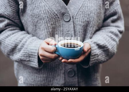 Eine Person in einem gemütlichen grauen Pullover hält eine blaue Tasse Kaffee und fängt einen ruhigen Moment der Wärme und des Komforts ein, perfekt für einen kühlen Tag. Stockfoto