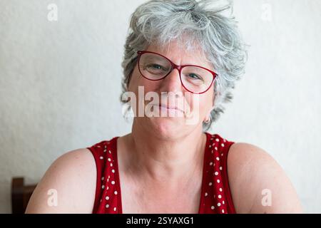 Eine lächelnde Seniorin mit kurzen grauen Haaren und roter Brille sitzt drinnen. Ihr herzlicher Ausdruck vermittelt Freundlichkeit und Freude Stockfoto