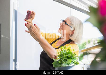 Eine ältere Frau mit Brille, gelbem Hemd und schwarzer Schürze hält ihr Smartphone hoch, um ein Foto zu machen. Sie steht auf einer hellen Balkonsurro Stockfoto
