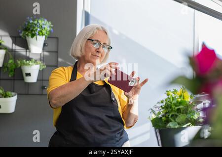 Eine ältere Frau mit weißem Haar und Brille, die ein gelbes Hemd und eine schwarze Schürze trägt, fotografiert mit einem Smartphone Blumen und Pflanzen in einem hellen i Stockfoto