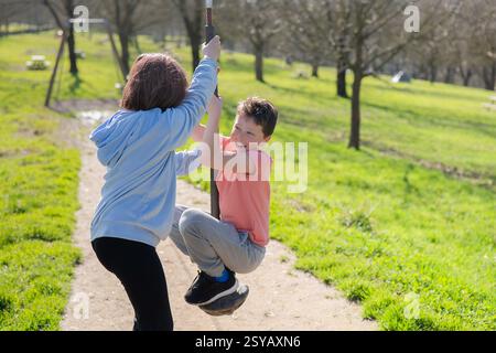 Zwei Freunde spielen fröhlich auf einer Seilrutsche in einem sonnigen Park. Umgeben von grünem Gras und Bäumen teilen sie Lachen und Freundschaft in der Natur Stockfoto