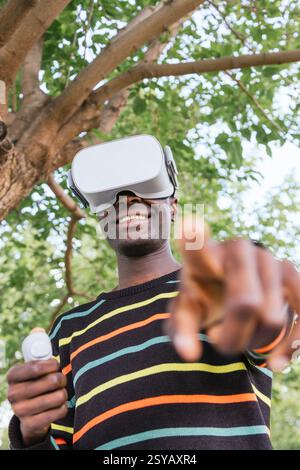 Ein lächelnder Mann erlebt die virtuelle Realität im Freien mit einer VR-Brille und einem gestreiften Pullover. Er zeigt auf die Kamera, hält einen VR-Controller, s Stockfoto