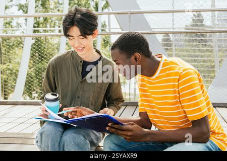 Zwei Schüler, die gemeinsam im Freien studieren, Notizen diskutieren und Ideen austauschen. Sie sitzen auf einer Holztreppe und genießen die Sonne und Akad Stockfoto