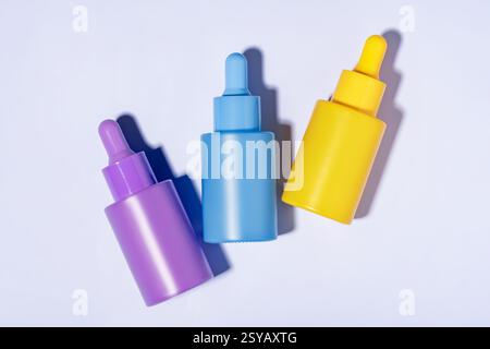 Draufsicht auf drei leuchtend farbige Kosmetikserumflaschen in violett, blau und gelb, ordentlich angeordnet auf einem sauberen weißen Hintergrund, die weiche Schatten bilden Stockfoto