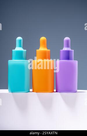 Drei leuchtende und farbenfrohe Serumflaschen, die auf einer schlanken Oberfläche mit einem weichen grauen Hintergrund ausgerichtet sind und die moderne Ästhetik der Hautpflege betonen Stockfoto