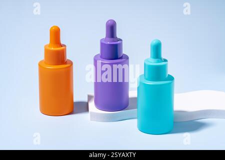Drei Serumflaschen in leuchtenden Orange-, Lila- und Türkisfarben, die auf einem sanften blauen Hintergrund präsentiert werden und moderne Kosmetikverpackungen demonstrieren Stockfoto