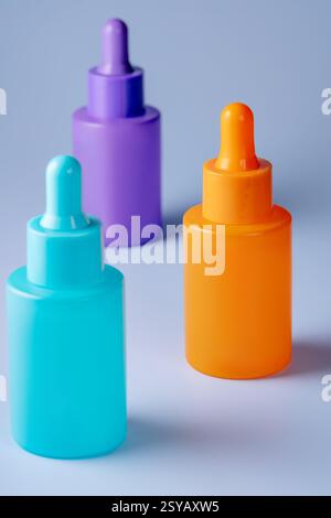 Drei leuchtend farbige Serumflaschen in Lila, Orange und Petrol, die ästhetisch auf einem hellgrauen Hintergrund angeordnet sind. Stockfoto