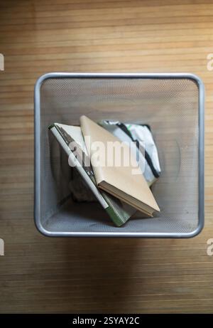 Luftaufnahme eines Abfallbehälters aus Metallgitter mit weggeworfenen Büchern auf Holzboden. Das Bild erinnert an Themen wie Abfall, Recycling und Umwelt Stockfoto