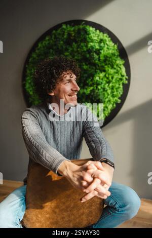 Ein junger Mann mit lockigen Haaren lächelt, während er an einer üppigen grünen Wand sitzt. Das Bild betont Themen der Ökologie und der nachhaltigen Entwicklung. Stockfoto