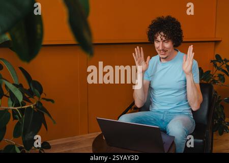 Ein junger Mann mit lockigen Haaren, der ein hellblaues Hemd trägt, beteiligt sich an einem Videoanruf auf seinem Laptop. Umgeben von Pflanzen und einem warmen orangen Hintergrund, er Stockfoto
