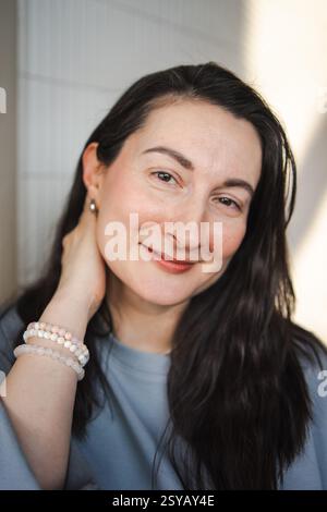Ein Porträt einer Frau mittleren Alters mit einem sanften Lächeln, die ein hellblaues Oberteil und Perlenarmbänder trägt und in sanftes natürliches Licht getaucht ist. Stockfoto