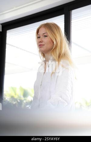 Eine junge Frau mit blonden Haaren, in einem weißen Hemd, steht in einem hellen, modernen Interieur. Große Fenster im Hintergrund ermöglichen natürliches Licht Stockfoto
