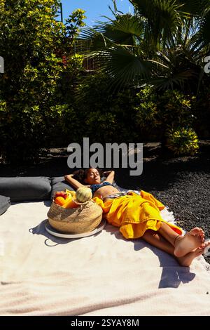 Ein Mädchen liegt auf einer Decke in einem Garten, sonnt sich unter klarem Himmel. Umgeben von üppigem Grün, entspannt sie sich mit einem Obstkorb in der Nähe, wo sie sich ertränkt Stockfoto