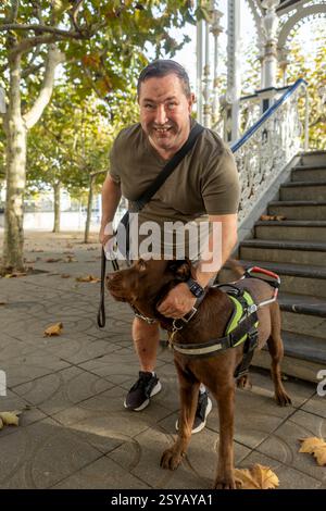 Ein lächelnder blinder Mann mit Blindenhund an der Leine genießt einen sonnigen Herbsttag in einem Park. Der Hund trägt ein Gurtzeug, und gefallene Blätter umgeben sie nahe dekorativ Stockfoto