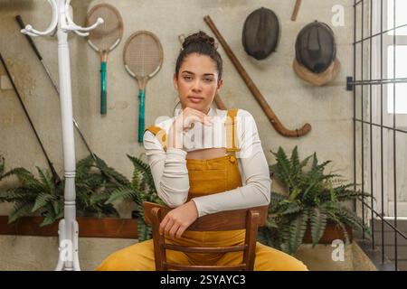 Eine junge Frau mit entspanntem Ausdruck, die in einem Holzstuhl sitzt, in gelben Overalls und einem weißen Hemd gekleidet, umgeben von Vintage-Sportausrüstung Stockfoto