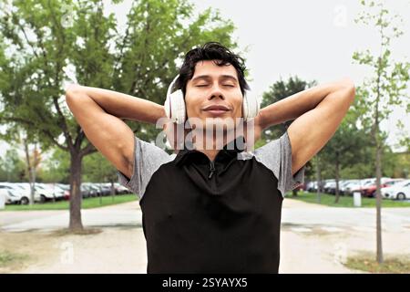 Ein männlicher Latino-Sportler genießt nach dem Training einen ruhigen Moment mit Kopfhörern an, steht draußen mit geschlossenen Augen und Bäumen im Hintergrund. Stockfoto