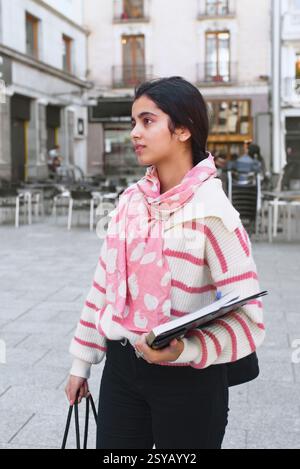 Eine arabische Studentin steht in einer Stadtstraße und hält ein Notizbuch und eine Tragetasche. Sie trug einen pink-weiß gestreiften Pullover und einen rosa Schal. Hintergrund Stockfoto