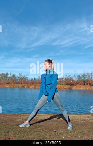 Eine Frau in Sportkleidung dehnt sich in der Nähe eines ruhigen Sees unter einem klaren blauen Himmel. Sie trägt eine blaue Jacke und Leggings und nimmt an einem Outdoor-Training Teil Stockfoto