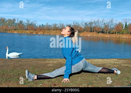 Eine Frau führt Yoga in einer malerischen Umgebung an einem ruhigen See auf. Sie dehnt sich in voller geteilter Haltung und trägt sportliche Ausrüstung. Ein Schwan gleitet auf der Wa Stockfoto