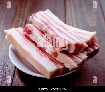 Die Scheiben von rohem Schweinebauch sind ordentlich auf einem weißen Teller auf einer rustikalen Holzoberfläche angeordnet. Das marmorierte Fett und das reichhaltige rote Fleisch betonen seine Fre Stockfoto