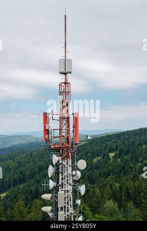 Ein hoch aufragendes Kommunikationsarray mit verschiedenen Antennen und Schüsseln, das vor dem Hintergrund von dichten, grünen Wäldern und fernen Hügeln steht Stockfoto
