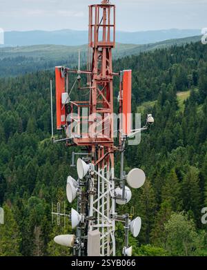 Detaillierte Ansicht eines Telekommunikationsturms mit Antennen und Funkschalen, sichtbar vor einem üppig grünen Waldgrund. Stockfoto