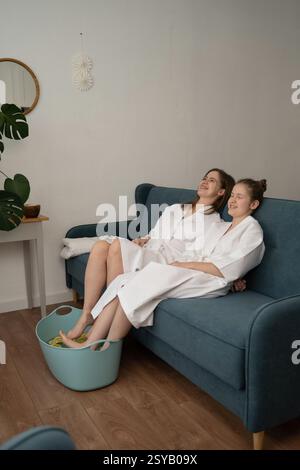 Mutter und Tochter genießen eine angenehme Zeit bei einem Spa-Besuch zu Hause. In weiße Bademäntel getaucht, tauchen sie ihre Füße in ein beruhigendes Zitronenfußbad, umarmt Stockfoto