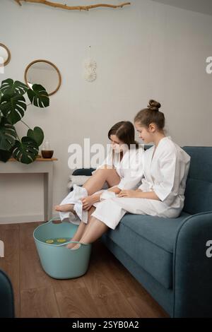 Mutter und Tochter verbringen gemeinsam einen entspannenden Heim-Spa-Tag, während sie ihre Füße in einer Badewanne mit Zitronenscheiben einweichen, während sie gemütliche weiße Bademäntel und Enj tragen Stockfoto