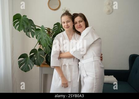 Mutter und Tochter in weißen Bademänteln teilen sich eine warme Umarmung während einer Selbstpflegesitzung zu Hause. Umgeben von Grün und ruhiger Einrichtung, reflektierend Stockfoto