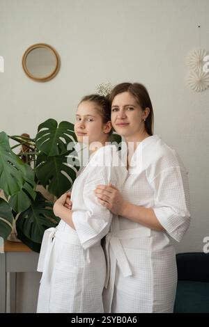 Mutter und Tochter verbringen zu Hause einen Tag der Selbstpflege. Sie tragen weiße Bademäntel, stehen zusammen und lächeln in einem gemütlichen Zimmer. Stockfoto