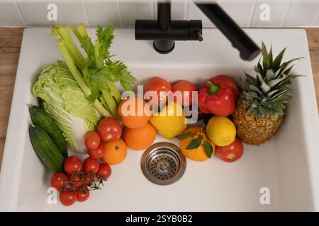 Eine Vielzahl von frischem Obst und Gemüse, einschließlich Tomaten, Orangen und Ananas, in einem weißen Spülbecken, bereit zum Waschen. Gesundes Essen Stockfoto