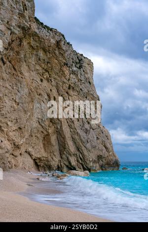 Der Strand von Porto Katsiki in Lefkada, Griechenland, besticht durch sein türkisfarbenes Wasser und seine zerklüfteten Klippen. Wellen umrunden sanft die Sandküste unter bewölktem Himmel. Stockfoto