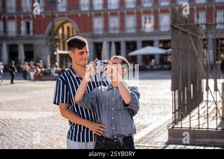 Ein fröhliches schwules Paar genießt einen sonnigen Tag in den belebten Straßen von Madrid. Mit einer Kamera fangen sie Erinnerungen auf, und sie umarmen Liebe und Glück in einer vibra Stockfoto