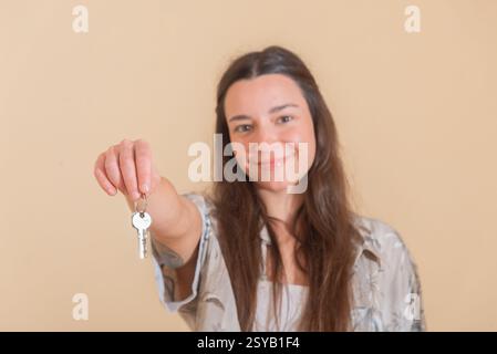 Eine lächelnde junge Frau hält mit einer einladenden Geste einen Schlüssel nach vorne und symbolisiert den Besitz eines Wohnhauses oder Immobilien im Umzug. Der warme Ton der Ba Stockfoto