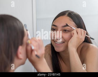 Eine Frau lächelt, während sie einen Augenbrauenstift vor einem Spiegel benutzt und sich darauf konzentriert, ihre Augenbrauen zu perfektionieren. Die Szene fängt das Wesen persönlicher Gentlemen ein Stockfoto