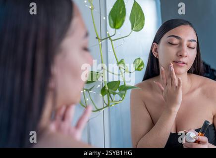 Eine Frau trägt Hautpflegecreme auf ihr Gesicht vor einem Spiegel auf. Sie konzentriert sich auf Selbstpflege, mit einer entspannten Atmosphäre und natürlichem Licht Stockfoto