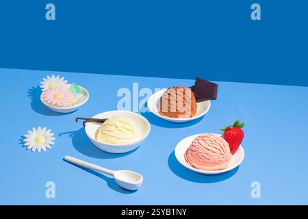 Drei verschiedene Eissorten auf einem leuchtend blauen Hintergrund, jeweils in einer weißen Schüssel mit dekorativen Elementen wie Erdbeere, Schokolade, o Stockfoto