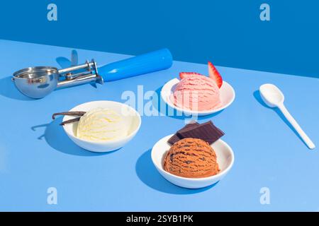 Drei Eissorten, Vanille, Erdbeere und Schokolade, serviert in weißen Schüsseln auf einem leuchtend blauen Hintergrund. Ein Löffel und ein weißer Löffel ergänzen Stockfoto