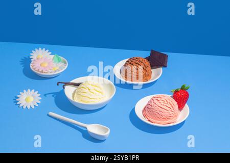 Bunte Eiskugeln in weißen Schüsseln unter einem kräftigen blauen Hintergrund, dekoriert mit Blumen, Schokolade und einer Erdbeere. Stockfoto