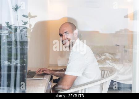 Lächelnder Mann, der in einem modernen Hotelzimmer an einem Laptop arbeitet. Natürliches Licht filtert durch das Fenster und schafft so eine ruhige Arbeitsatmosphäre, die sich positiv auf die Arbeit auswirkt Stockfoto