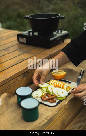Zugeschnittene, nicht erkennbare Person, die Zutaten für Glühwein im Freien zubereitet; geschnittene Äpfel und Orangen auf einem Holztisch anordnen. Stockfoto