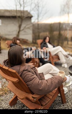 Drei unbekannte Frauen genießen ein entspanntes Treffen im Freien, sitzen auf Holzstühlen und trinken warme Getränke in einer ruhigen, natürlichen Umgebung. Der Fokus Stockfoto